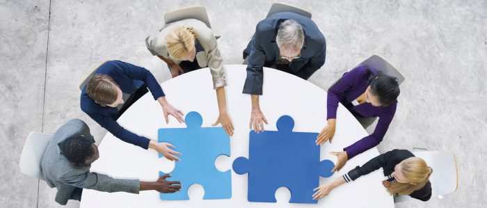Sechs Menschen, die gemeinsam um einen Tisch herum sitzen, fügen zwei Teile eines Puzzles zusammen.