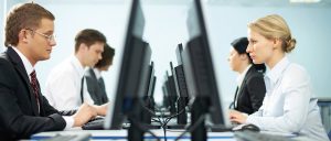 On the left side, two men are sitting in front of their desktop. On the richt side, two women are sittng in front of their desktops, opposite to the men. The picture is a symbol of the gender wage gap.