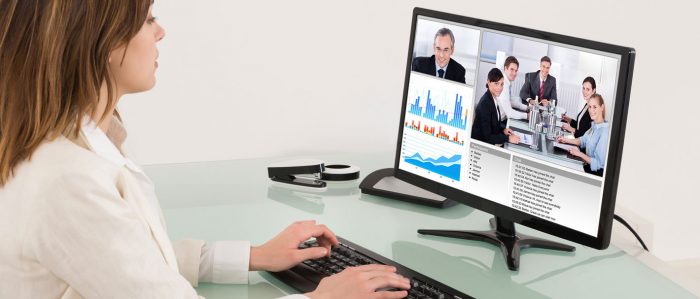 The picutre shows a woman sitting at a desk, taking part in a video conferen that she watches on her PC screen while typing on the keyboard.