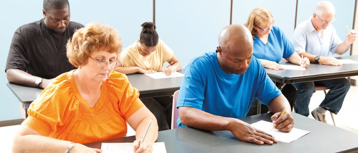 Sechs Menschen mit unterschiedlicher Herkunft, Alter und Geschlecht sitzen in einem Raum und schreiben eine Prüfung