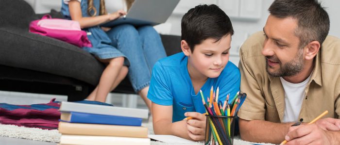 Mann mit Sohn liegen vor Hausaufgaben auf dem Boden. Im Hintergrund sitzt die Mutter mit der Tochter auf dem Bett, Laptop auf dem Schoß.