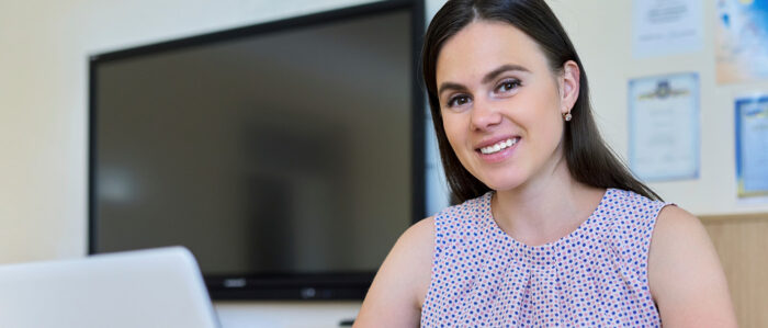 A young woman with dark hair and dark eyes sits, wearing a sleeveless blouse sits at a desk in an office.