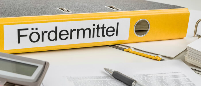 Gelber Ordner mit der Aufschrift „Fördermittel“ liegt auf einem Schreibtisch