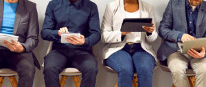 Drei Männer und eine Frau sitzen nebeneinander in einem Wartezimmer und arbeiten an ihren Laptops.