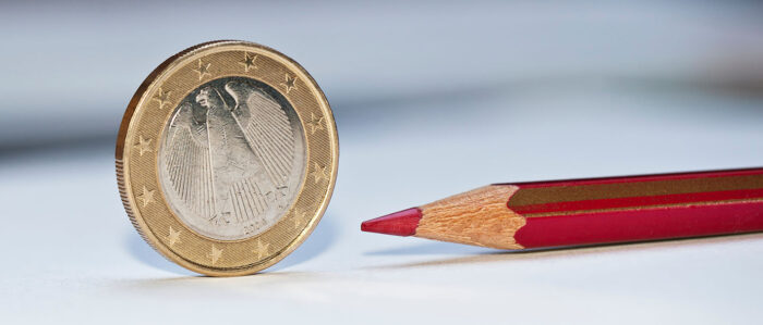 Eine Ein-Euro-Münze steht hochkant auf einer Fläche, daneben liegt ein Rotstift.