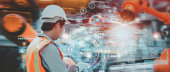 Industriearbeiter in mit Warnweste und Helm von hinten, der über ein Tablet Roboterarme steuert.