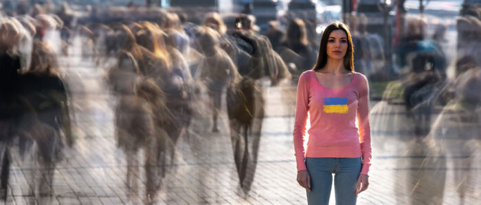 Junge Frau mit einer ukrainischen Flagge auf dem T-Shirt steht in einer Fußgängerzone und schaut den Betrachter mit leerem Blick an. Um sie herum laufen Menschen.