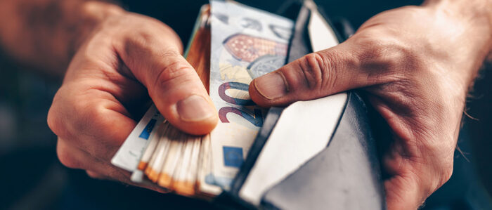 Nahaufnahme der Hände eines Mannes, die einen geöffneten Geldbeutel halten und einen aufgefächerten Stapel Geldscheinen, der zur Hälfte darin steckt.