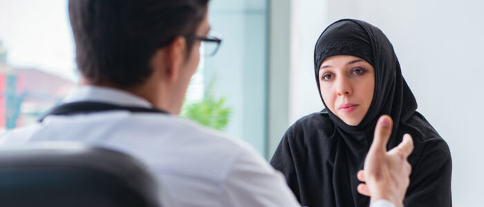 Das Bild zeigt eine Beratungssituation. Der Mann, der von hinten abgebildet ist, erklärt einer Frau mit Kopftuch, vermutlich aus dem Nahen Osten, etwas, die auf der gegenüberliegenden Seite des Tisches sitzt und ihm zuhört.
