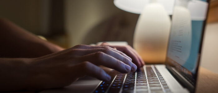 Nahaufnahme von Händen, die im Licht einer Tischlampe auf der Tastatur eines Laptops tippen.