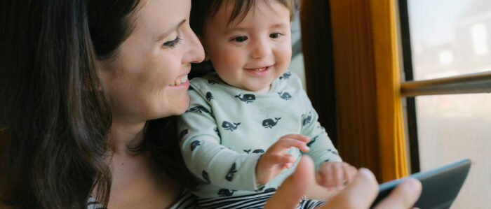 Eine Mutter schaut ihr Kleinkind an, das sie auf dem Arm hat, und zeigt ihm etwas auf einem Tablet. Beide lächeln und sehen glücklich aus.
