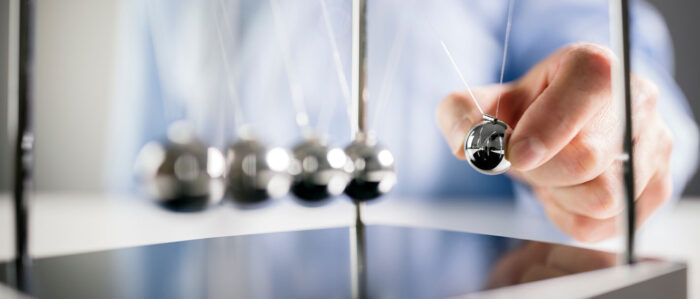 Das Bild zeigt eine männliche Hand, die die äußere Kugel eines Newtonpendels wegzieht um dieses anzustoßen.