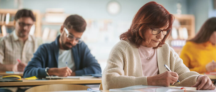 Ältere Teilnehmerin einer Fortbildung sitzt in einem Kursraum an ihrem Schreibtisch und notiert sich etwas.