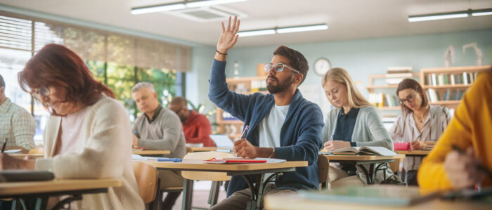 Frauen und Männer sitzen an Tischen, angeordnet wie in einer Schule. Ein Mann hebt seine Hand, um sich zu melden.