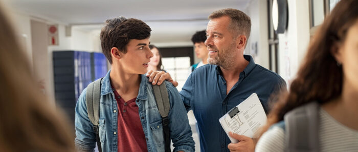 Lehrer und sein Schüler unterhalten sich während sie über den Gang im Schulgebäude laufen. Der Lehrer legt dem Schüler väterlich die Hand auf die Schulter.