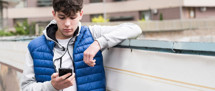 Ein Jugendlicher steht vor einem Wohnblock und schaut auf sein Handy, das er in der Hand hält.