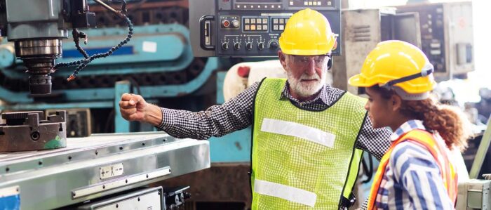 Ein älterer grauhaariger Arbeiter erklärt einer jungen Arbeiterin die Funktion einer Metalldrehmaschine.