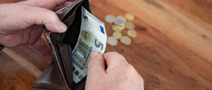 Nahaufnahme: Ein Mann hält seinen Geldbeutel in der einen Hand und zieht mit der anderen den letzten 5-Euro-Schein heraus. Im Hintergrund liegen einige Münzen auf dem Tisch