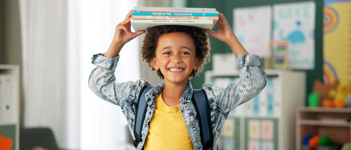 Lachendes Kind hält den Stapel Bücher auf seinem Kopf mit beiden Händen fest.