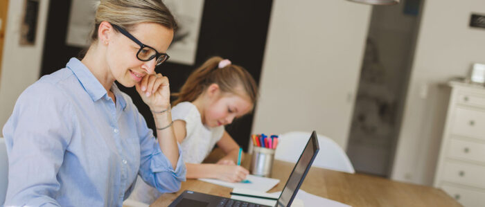 Junge Mutter sitzt zu Hause am Tisch und arbeitet an einem Laptop. Dahinter am selben Tisch sitzt Ihre kleine Tochter und malt.