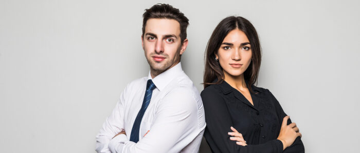 Junge Frau und junger Mann, beide in legerer Business-Kleidung, stehen Schulter an Schulter vor einem grauen Hintergrund und schauen in die Kamera.