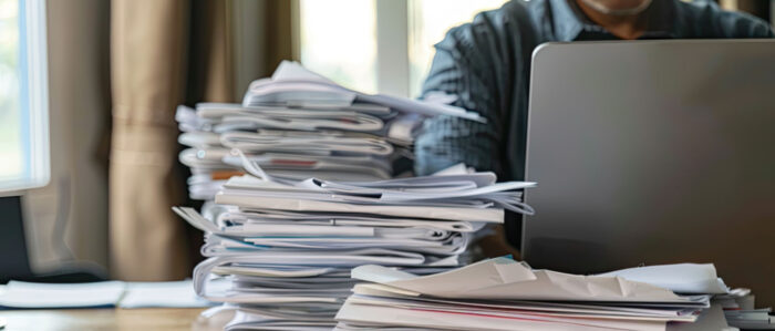 Eine Person sitzt am Schreibtisch und arbeitet am Laptop. Daneben stapeln sich Dokumente.