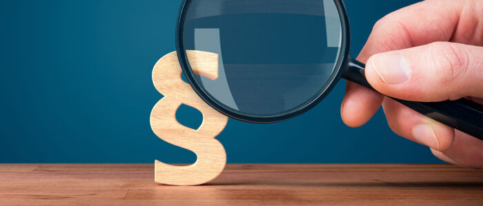 Auf den Bild ist ein Paragraphenzeichen zu sehen, das von einer großen Hand mit einer Lupe betrachtet wird.