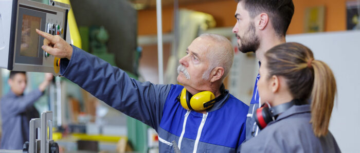 Meister erklärt seinen zwei Lehrlingen etwas an einer Maschine.