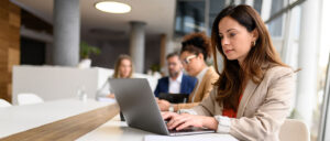 Eine junge Geschäftsfrau schreibt in einem Sitzungssaal konzentriert mit ihrem Laptop. Im Hintergrund arbeiten ihre Kollegen.