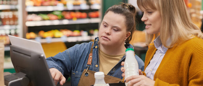 Porträt einer jungen Frau mit Down-Syndrom, die im Supermarkt an einer Selbstbedienungskasse einer Kundin hilft.