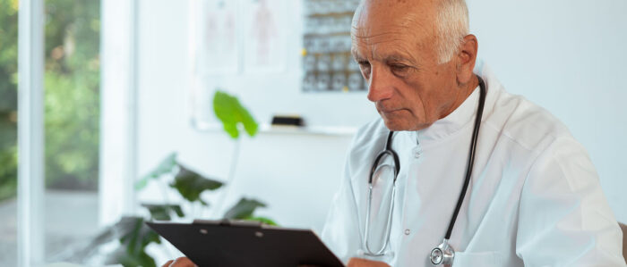 Älterer Arzt im weißen Kittel und mit Stethoskop um den Hals sitzt am Schreibtisch und schaut auf ein Klemmbrett.