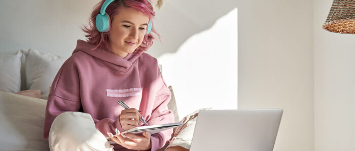 Jugendliche mit rosa Haaren und türkisen Kopfhörer sitzt lächelnd auf Ihrem Bett vor ihrem Laptop und macht sich Notizen.