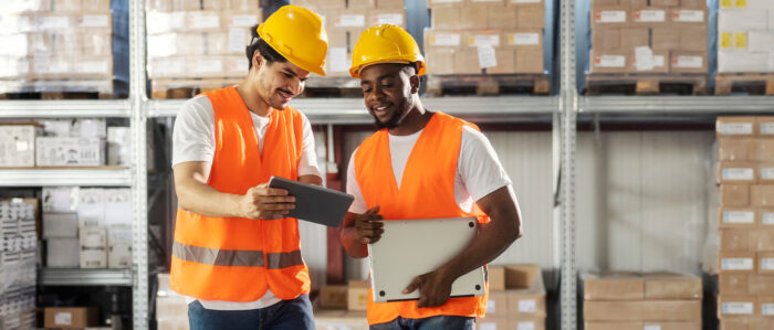 Zwei ausländische Lageristen stehen in einem Warenlager, und sprechen lächelnd über etwas, dass sie auf dem Tablet sehen, das der eine in der Hand hält.