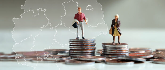 Zwei Miniatur-Figuren, ein Mann und eine Frau, stehen auf je einem Münzstapel, wobei der Münzstapel des Mannes höher ist, als der der Frau. Im Hintergrund ist eine Landkarte mit dem Umriss von Deutschland zu sehen.